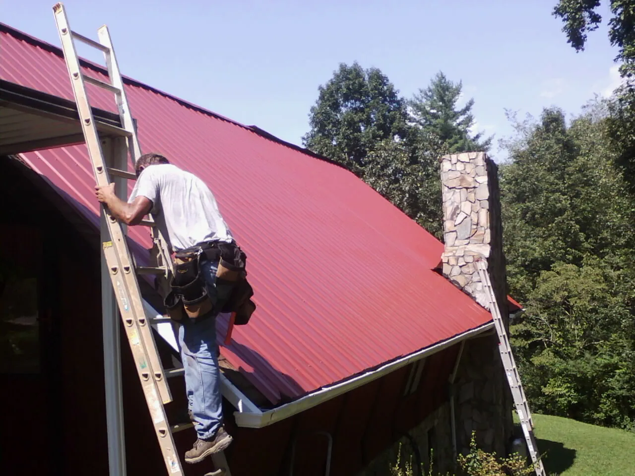 Roofer on a ladder beside a red metal roof and stone chimney — Soffit & Fascia Repair by Roofing Brothers in Beyer